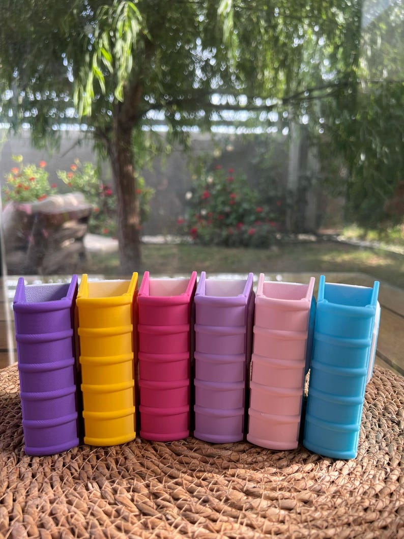 Puede incluir: Seis peque&ntilde;as macetas con forma de libro, de colores vibrantes: morado, amarillo, rosa, lavanda, rosa claro y azul. Las macetas est&aacute;n dispuestas en fila sobre una superficie tejida.