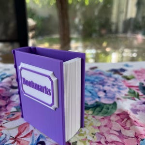 Puede incluir: Un soporte para marcap&aacute;ginas morado con forma de libro, con la palabra "Bookmarks" en letras blancas en la parte delantera. El soporte est&aacute; sobre una superficie con estampado floral. Las p&aacute;ginas son blancas.