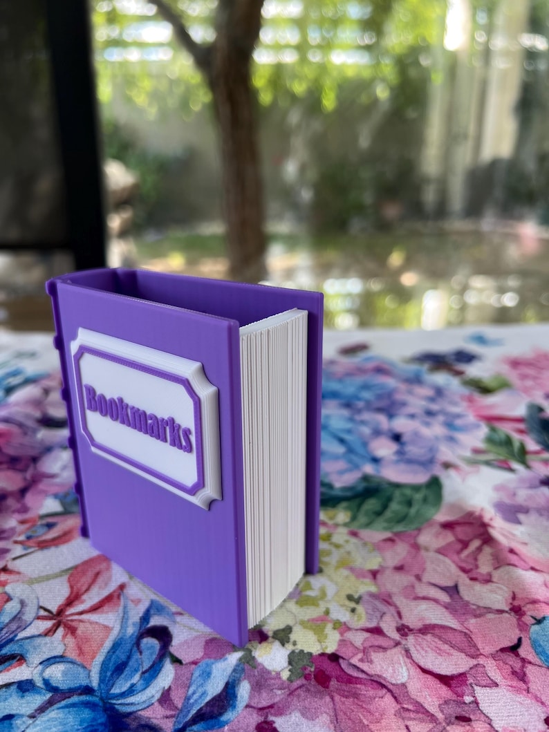 Puede incluir: Un soporte para marcap&aacute;ginas morado con forma de libro, con la palabra "Bookmarks" en letras blancas en la parte delantera. El soporte est&aacute; sobre una superficie con estampado floral. Las p&aacute;ginas son blancas.