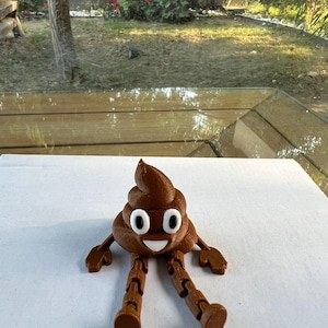 Puede incluir: Figura de emoji de caca marr&oacute;n impresa en 3D con ojos blancos y boca sonriente. La figura tiene brazos y piernas articulados y est&aacute; colocada sobre una superficie blanca. El fondo muestra una escena exterior borrosa con vegetaci&oacute;n.