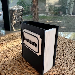 Puede incluir: Un contenedor negro con forma de libro con la palabra "Bookmarks" en letras blancas sobre una etiqueta blanca y negra. El contenedor est&aacute; lleno de papel blanco y descansa sobre una superficie tejida.