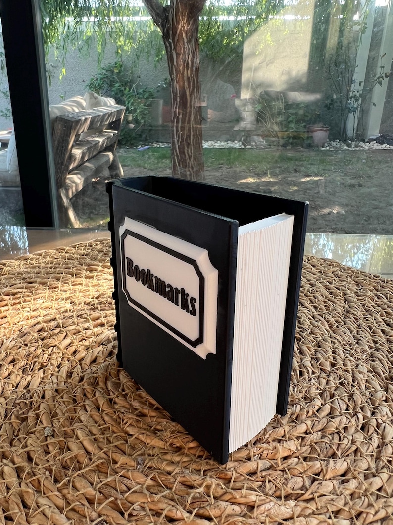Puede incluir: Un contenedor negro con forma de libro con la palabra "Bookmarks" en letras blancas sobre una etiqueta blanca y negra. El contenedor est&aacute; lleno de papel blanco y descansa sobre una superficie tejida.