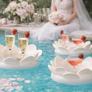 May include: White lotus-shaped floating drink holders in a pool, each holding a champagne glass with a strawberry garnish. The water is a clear blue, and a bride in a wedding dress is in the background.