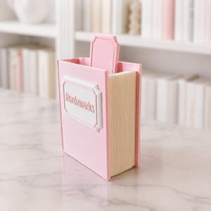 Puede incluir: Un organizador de escritorio rosa con forma de libro con la palabra "Bookmarks" en una etiqueta blanca. El organizador tiene una ranura para guardar objetos y una pesta&ntilde;a decorativa. El lateral del organizador se asemeja a p&aacute;ginas de libro.