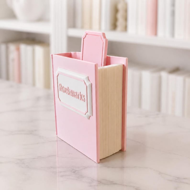 Puede incluir: Un organizador de escritorio rosa con forma de libro con la palabra "Bookmarks" en una etiqueta blanca. El organizador tiene una ranura para guardar objetos y una pesta&ntilde;a decorativa. El lateral del organizador se asemeja a p&aacute;ginas de libro.