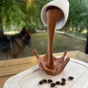 May include: A white ceramic coffee cup with brown liquid pouring out of it, creating a splash. The liquid is frozen in mid-air, and there are coffee beans scattered on the white surface below.