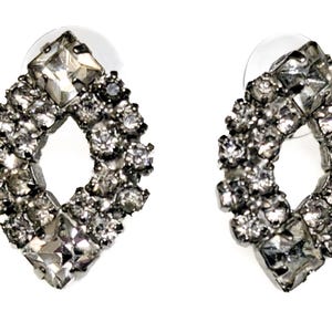 Puede incluir: Un par de pendientes de botón plateados en forma de diamante. Cada pendiente presenta una piedra central grande, de talla cuadrada, rodeada de piedras redondas más pequeñas. Los pendientes están sobre un fondo blanco.