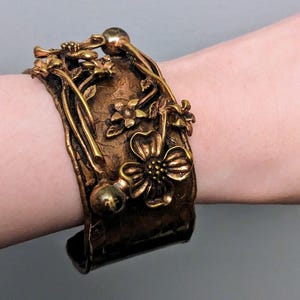 May include: A wide, hammered copper bracelet with a floral design. The bracelet is adorned with raised flowers and leaves, and has a textured surface. The bracelet is a warm, golden brown color.