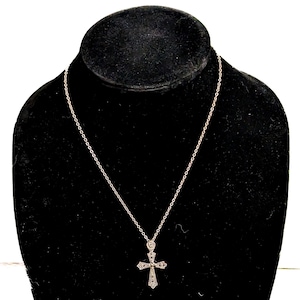 Könnte beinhalten: Eine silberfarbene Halskette mit einem Kreuzanhänger. Das Kreuz ist mit kleinen, funkelnden Akzenten versehen. Die Halskette wird auf einer schwarzen Samtbüste präsentiert, die das Design und die Handwerkskunst des Schmucks hervorhebt.