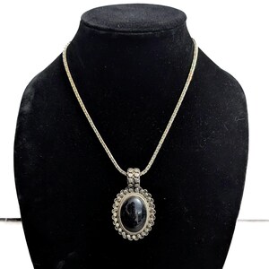 May include: A silver-toned necklace with a large, oval black stone pendant. The pendant is surrounded by a decorative border and hangs from a delicate chain. The necklace is displayed on a black mannequin.