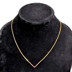 Puede incluir: Un collar dorado con un colgante en forma de V engastado con pequeñas piedras rojas. El collar se exhibe sobre un busto de joyería de terciopelo negro, mostrando su delicada cadena y vibrante colgante.