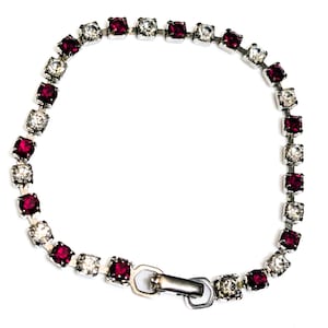 Puede incluir: Una pulsera plateada con piedras cuadradas rojas y transparentes alternas. La pulsera tiene un cierre sencillo y se muestra sobre un fondo blanco. Esta joya es adecuada para diversas ocasiones.