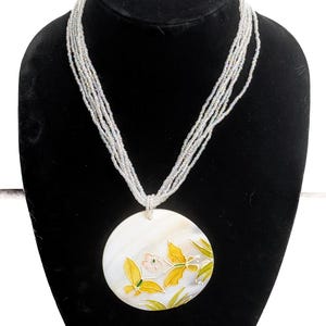 Puede incluir: Un collar de cuentas blancas con un colgante grande y redondo. El colgante presenta un diseño floral con hojas amarillas y verdes y una flor rosa. El collar se exhibe en un maniquí negro.