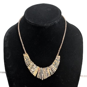 May include: A necklace with a curved pendant made of rectangular, patterned stones in shades of beige, brown, and gray. The necklace is displayed on a black mannequin. The necklace's chain is a thin, dark brown.