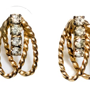 May include: A pair of gold-coloured earrings with a twisted rope design. Each earring has three clear, sparkling stones set vertically in the centre. The earrings are set against a white background.