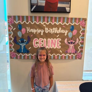 May include: A birthday banner with the words "Happy Birthday CELINE" in white script, featuring cartoon characters and balloons. A young girl wearing pink glasses and a pink shirt stands in front of the banner.