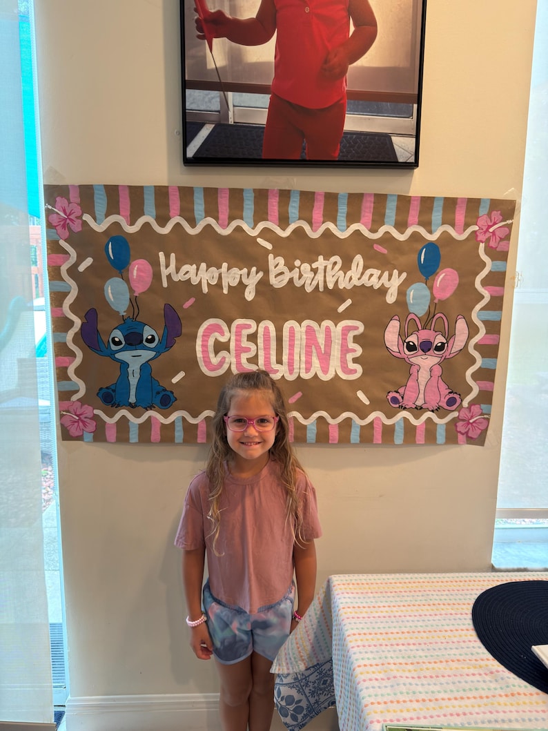 May include: A birthday banner with the words "Happy Birthday CELINE" in white script, featuring cartoon characters and balloons. A young girl wearing pink glasses and a pink shirt stands in front of the banner.