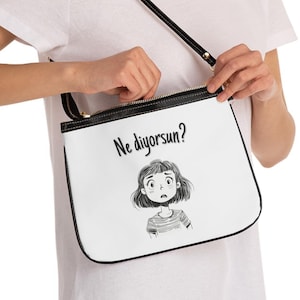 Könnte beinhalten: Weiße Umhängetasche mit schwarzem Trageriemen und schwarzem Besatz. Die Tasche hat eine Comic-Illustration eines Mädchens mit dem Text "Ne diyorsun?" darauf gedruckt.