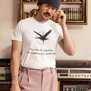 May include: A white t-shirt with a black albatross graphic and the text "You are my albatross, my compass in a stormy sea". The person is wearing a grey beanie and glasses.