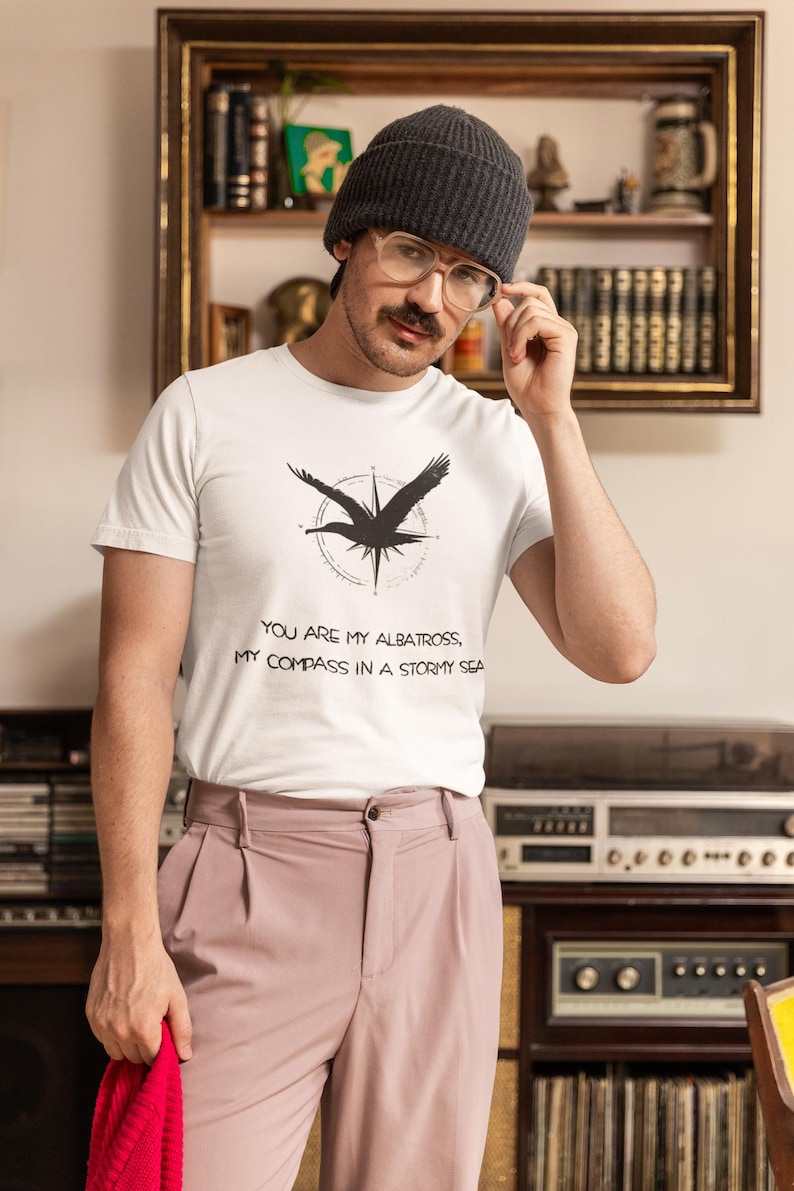 May include: A white t-shirt with a black albatross graphic and the text "You are my albatross, my compass in a stormy sea". The person is wearing a grey beanie and glasses.