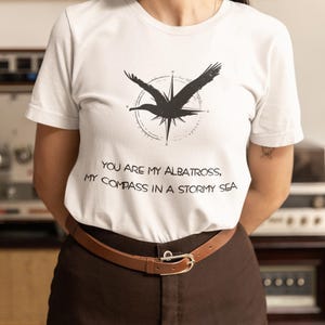 May include: White t-shirt with a black silhouette of an albatross flying over a compass. The text "You are my albatross, my compass in a stormy sea" is printed on the shirt.