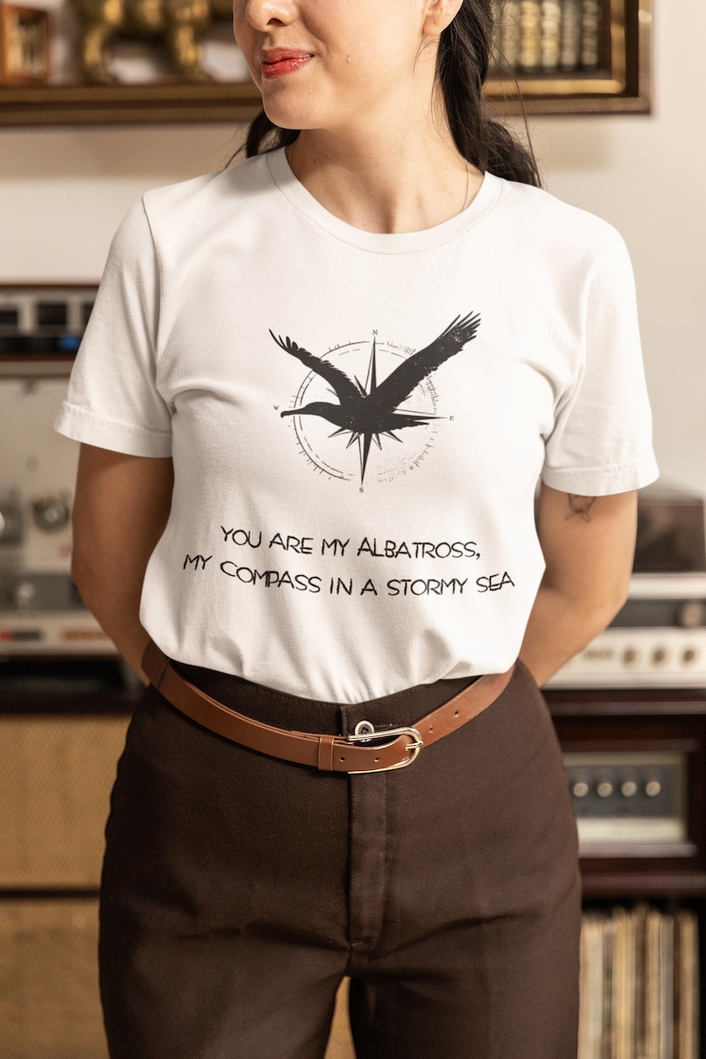 May include: White t-shirt with a black silhouette of an albatross flying over a compass. The text "You are my albatross, my compass in a stormy sea" is printed on the shirt.
