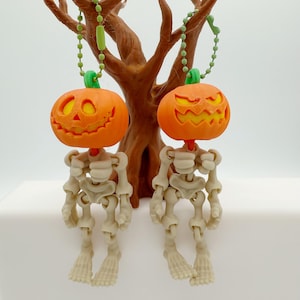 May include: Two orange pumpkin-shaped Halloween ornaments with carved faces hang from green beaded strings. Each pumpkin is attached to a skeleton figure with articulated limbs. A brown tree-like structure serves as a backdrop.