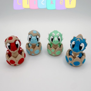 May include: Four small dinosaur figurines emerging from cracked eggs, arranged in a row. The text "clicker" appears in colorful pastel blocks above the figures. Each dinosaur has a distinct color scheme: red, light blue, green, and dark blue.