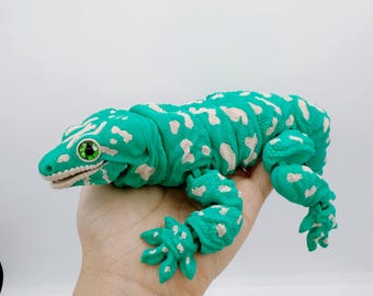 May include: A green and white spotted gecko toy with articulated limbs and a detailed head. The gecko has a bright green body with white spots and green eyes. The toy is held in a hand against a white background.
