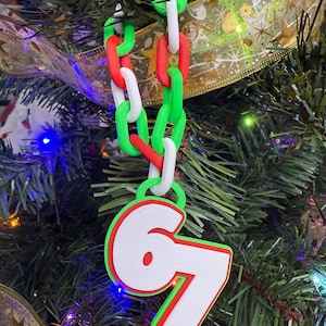 May include: A Christmas tree decoration with a chain of red, white, and green links. The ornament displays the number "67" in white with a red outline and a green border. It is suspended from a Christmas tree.