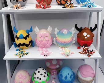May include: A collection of twelve decorative eggs and twelve small articulated dragon figurines displayed on a three-tiered white shelf. The eggs feature various patterns, colors, and themes, including floral, spotted, and creature-inspired designs.