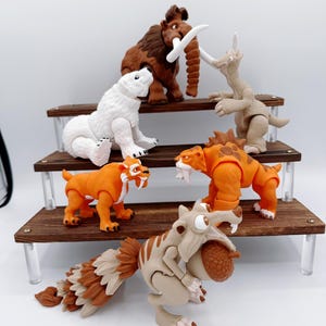 May include: A collection of Ice Age movie character figurines, including a brown mammoth, a white polar bear, and orange saber-tooth tigers, displayed on a multi-tiered wooden shelf. The figures are detailed and colorful, with a focus on the characters' distinctive features.