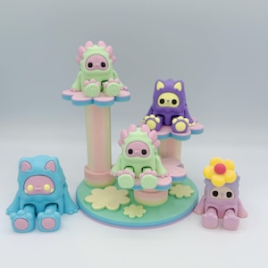 May include: A collection of colorful, whimsical figurines. The figures are in the shape of small creatures wearing hooded outfits, some with flower-shaped hats. They are in shades of blue, green, purple, and pink, with pastel-colored platforms.