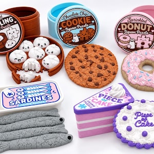 Puede incluir: Una colección de rompecabezas con temática de comida que incluye una vaporera de dumplings marrón, una lata azul de galletas con chispas de chocolate, una caja de donas rosa, una lata de sardinas con piezas de peces grises y dos pasteles.
