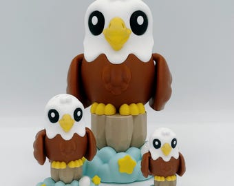May include: Three decorative eagle figurines. The largest eagle is brown and white, perched on a wooden base. Two smaller eagles rest on blue cloud bases with yellow stars. The eagles have large black eyes and yellow beaks.
