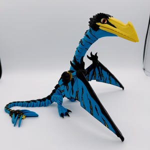 May include: A blue and black articulated dinosaur figurine with a long neck and a prominent yellow beak. The creature features black wing membranes with blue accents, sharp claws on its limbs, and a long tail ending in a small yellow cross-shaped detail.