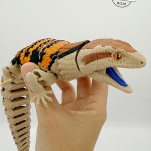 May include: A detailed, articulated lizard toy with a tan body, black and orange markings, and a brown head. The lizard's mouth is open, revealing a blue tongue. The toy is held in a hand, with the "AUTHORIZED RAKI-BOX SELLER" logo visible.