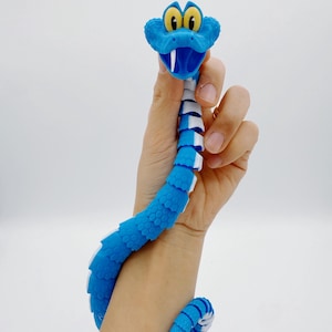Op de afbeelding: Een blauw-wit gearticuleerd slangen speelgoed met gele ogen en een witte tong. Het speelgoed wordt door een hand omhoog gehouden, met het lichaam van de slang om de pols gewikkeld. De kop van de slang is omhoog en het lichaam is gesegmenteerd.