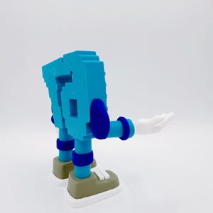 May include: A 3D-printed, pixelated figure in shades of blue, with a white hand extended. The figure has a blocky design, with a light blue body, dark blue accents, and green and white shoes. The figure is standing.