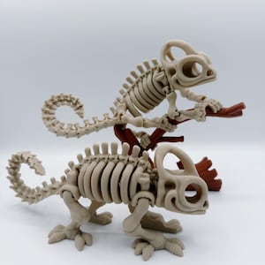 May include: Two beige chameleon skeleton figurines, one perched on a brown branch. The skeletons have detailed rib cages, leg bones, and tail structures. The background is plain white.