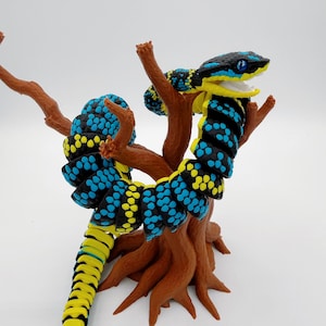 Könnte beinhalten: Eine mehrgliedrige, bewegliche Schlangenfigur mit schwarzem Körper und leuchtend blauen und gelben Schuppenmustern. Die Schlange ist um einen braunen, strukturierten, baumartigen Ständer gewickelt. Sie hat ein weit geöffnetes Maul und leuchtend blaue Augen.