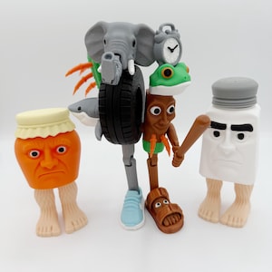 Op de afbeelding: Een grillige sculptuur met antropomorfe figuren. Een oranje pot met een gezicht en voeten staat naast een meerdelige figuur met een olifantenkop, bandenlichaam en klok. Een zoutstrooier met een gezicht en voeten houdt een honkbalknuppel vast.
