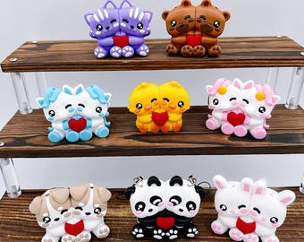 Puede incluir: Una colección de figuras de animales coloridas y emparejadas, que incluyen gatos, osos, perros, pandas y conejos. Cada figura está conectada y sostiene un corazón rojo. Las figuras se exhiben en una estantería de madera de tres niveles.