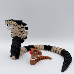May include: Two articulated snake toys. The larger snake has a black and tan head and body, with a black tail. The smaller snake is brown with blue eyes and a red tongue. Both snakes are on a white background.