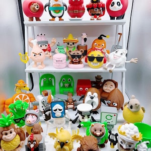 May include: A display of quirky figurines on white shelves. The figurines represent a variety of characters, including animals, fruits, and food items, each with distinct features and accessories. The collection includes a bear in an apple, a penguin, and a panda.