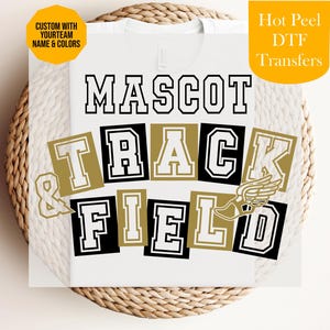 Könnte beinhalten: Ein weißes T-Shirt mit einem Grafikdesign, das den Text MASCOT TRACK & FIELD in einer College-Schriftart zeigt. Die Buchstaben sind weiß mit schwarzen und goldenen Umrandungen und in Blöcken angeordnet. Ein geflügeltes Schuhsymbol ist integriert.