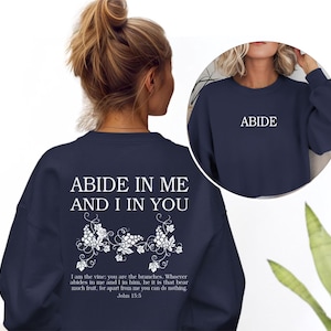 May include: A navy blue long-sleeve sweatshirt featuring white text and a grape vine graphic on the back. The back displays the text ABIDE IN ME AND I IN YOU above a vine illustration and the verse John 15:5. The front features the word ABIDE.