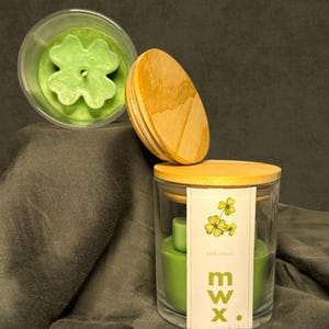 May include: A clear glass jar candle with a wooden lid, and a four-leaf clover-shaped candle. The candle is green and has a label that says "Irish Cream" and "mw x."