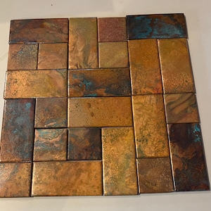 May include: A mosaic of rectangular copper tiles arranged in a brick pattern. The tiles display a range of colours, including shades of brown, gold, and teal, with a textured, weathered appearance. The tiles are arranged in a square shape.