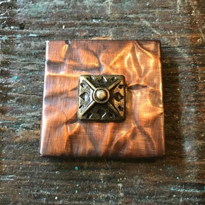 May include: A square copper metal decorative piece with a hammered texture and a brass-colored square embellishment in the center. The embellishment has a cross design with a central circular element. The piece is set on a wooden surface.